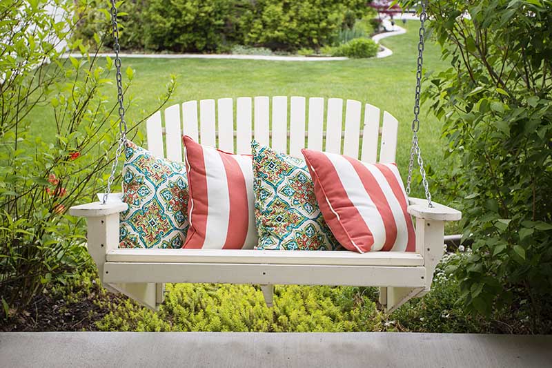 A close up of a white hanging bench overlooking a summer garden scene with lawns and shrubs in the background.