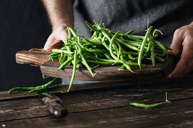 A close up of a man holding a wooden chopping board with a fresh harvest of green bush beans and a knife in the foreground.