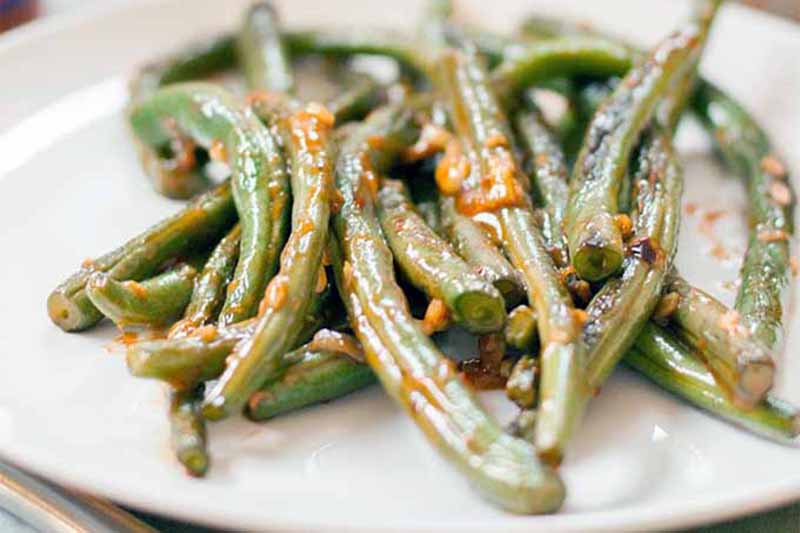 A close up of a white plate with garlic roasted green beans on a soft focus background.