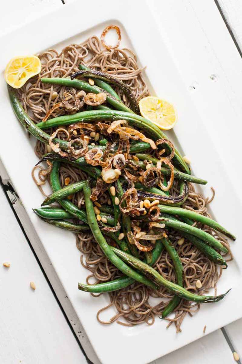 A close up vertical picture of a rectangular white plate with green beans and soba noodles topped with pine nuts and caramelized onion. To the side of the plate is lemon sections.