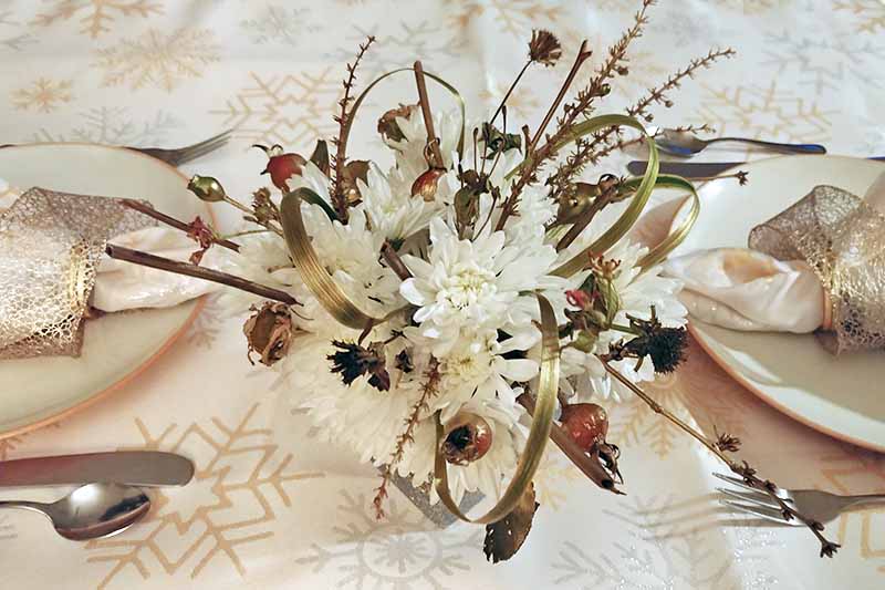 Overhead shot of a centerpiece comprised of white chrysanthemums, grasses, rose hips, and twigs from the garden, at the center of a table set with two white and orange plates with cream-colored cloth napkins tied with gold ribbon on top, with silverware at each place setting, on a cream-colored tablecloth with a gold and silver snowflake pattern.