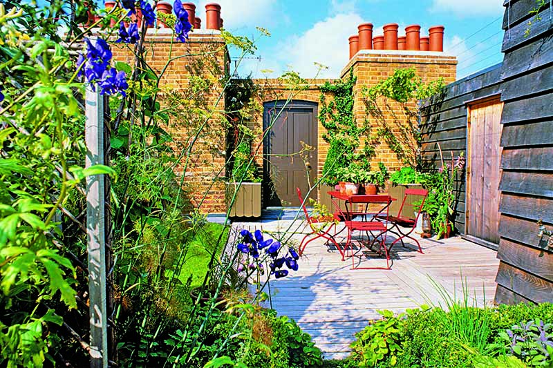 A rooftop deck with a table and chairs, and various plants growing around the border, with a brick chimney topped with red chimney pots, and a blue sky in the background.
