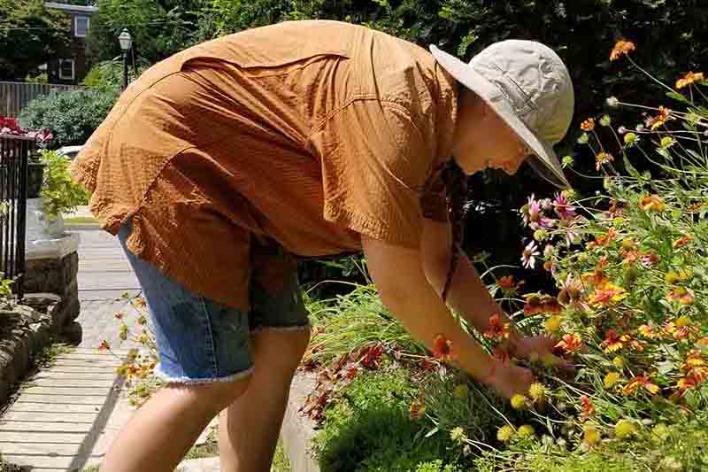 A woman weeding a garden, wearing a short sleeved sun protective blouse, and a wide brimmed hat with blue sky and trees in the background.