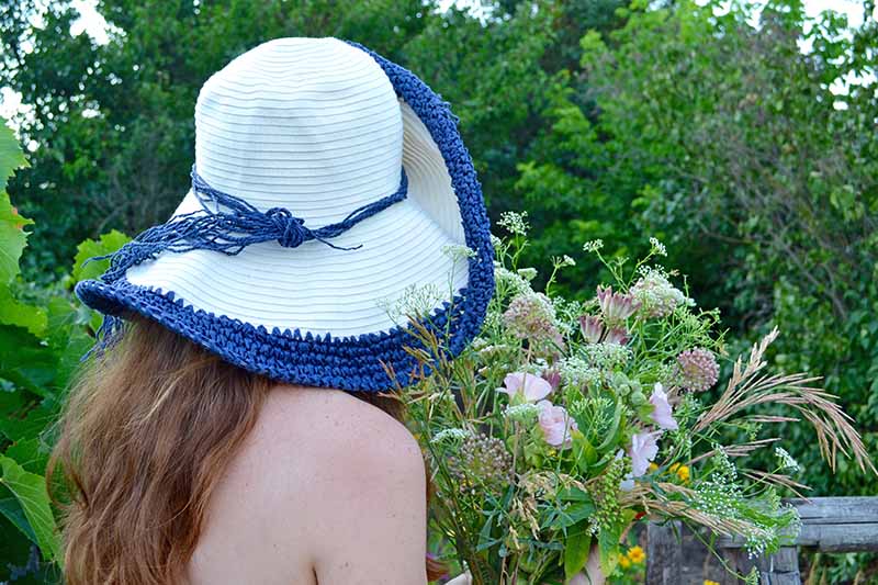 A close up of a young woman with bare shoulders wearing a blue and white hat holding a bunch of freshly picked flowers with trees in soft focus in the background.